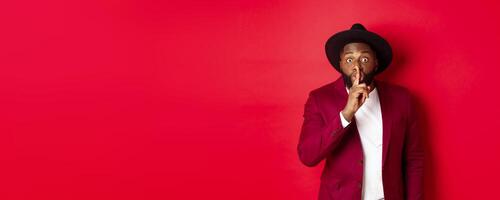 Fashion and party concept. African american man in classy hat hushing, asking keep secret, preparing surprise, standing over red background photo