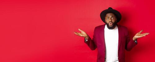 Confused and worried Black man spread hands sideways, staring at camera puzzled, asking what wrong, standing against red background photo