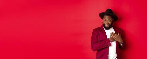 Fashion and party concept. Handsome african american macho man flirting, smiling and holding hands on heart, being grateful and touched, standing over red background photo