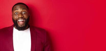 Close-up of african american man with beard staring amazed at camera, smiling and looking excited, standing in blazer against red background photo