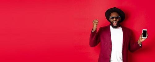 Cheerful winning Black man showing smartphone screen, rejoicing and demonstrating application, standing over red background photo