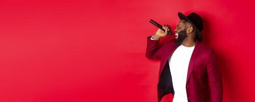 Passionate black male singer performing against red background, singing into microphone, wearing party outfit, standing over red background photo