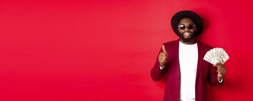 Handsome modern african american man in sunglasses and party clothes, showing thumb up with dollars, earn money and looking satisfied, red background photo