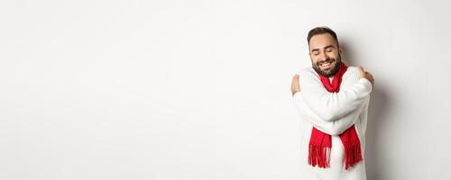 Happy bearded man enjoying comfortable winter sweater, smiling with warmth, hugging himself, standing over white background photo