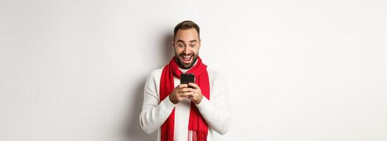 Man reading messaging on mobile phone and looking happy, standing in winter sweater and red scarf, white background photo