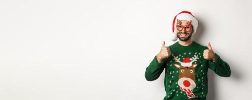 Christmas holidays, celebration concept. Happy man in Santa hat and party glasses showing thumbs up in approval, like something, standing over white background photo