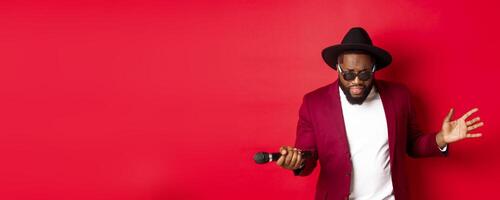 Passionate black male singer performing against red background, singing into microphone, wearing party outfit, standing over red background photo