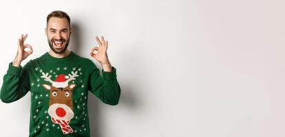 Christmas, holidays and celebration. Satisfied handsome guy in sweater, showing okay signs and smiling, praise something great, standing over white background photo