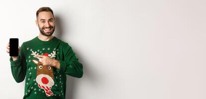New Year, holidays and celebration. Handsome man smiling, pointing finger at mobile screen, showing app, standing over white background photo