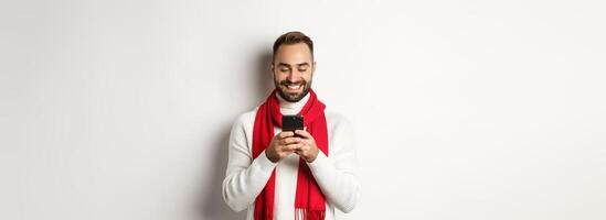 Happy man smiling while reading message on mobile phone, standing in winter sweater and red scarf against white background photo