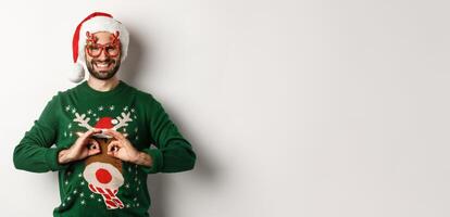 Christmas holidays, celebration concept. Happy guy in Santa hat and party glasses making fun of funny sweater, standing over white background photo