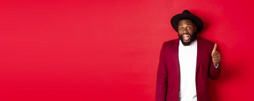 Christmas shopping and people concept. Handsome bearded Black man in party blazer showing thumb up, looking amazed and satisfied, red background photo