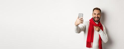 Handsome guy wishing merry christmas on call, waving hand at mobile phone and smiling, standing in sweater with red scarf, white background photo