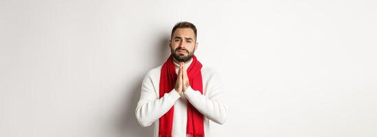 Christmas holidays and New Year concept. Desperate man begging for help, asking for favour, holding hands in pray and looking with hope at camera, standing over white background photo
