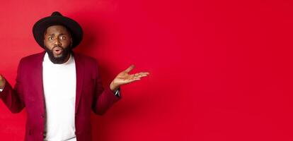 Confused and worried Black man spread hands sideways, staring at camera puzzled, asking what wrong, standing against red background photo