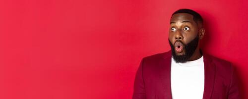 Close-up of impressed Black man looking left at copy space, saying wow and staring with amazement, standing over red background photo