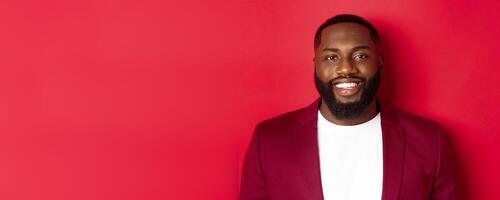 Close-up of handsome and stylish Black man wearing party blazer, smiling happy at camera, standing against red background photo