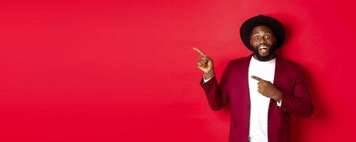 Winter holidays and shopping concept. Happy Black man pointing left and smiling, showing new year promo offer on red background photo