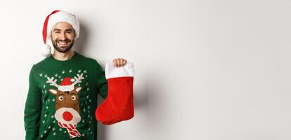 Xmas party and holidays concept. Happy man in Santa hat bringing gifts in Christmas sock and smiling, standing over white background photo