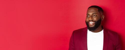 Close-up of handsome african american man with beard, looking left and smiling joyful, standing over red background photo