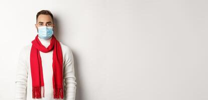 Covid-19, lockdown and quarantine concept. Young man in winter sweater and medical mask standing against white background photo