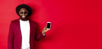 Handsome and stylish Black man showing phone screen at camera, recommending online store or application, standing over red background photo