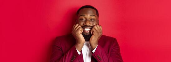 Close-up of silly african american man watching something adorable, touching cheeks and smiling with delight and happiness, standing over red background photo