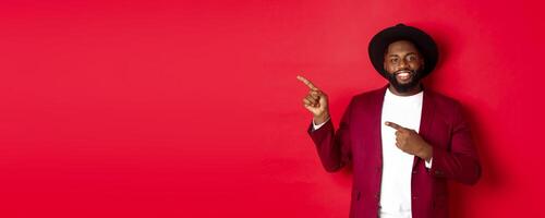 Winter holidays and shopping concept. Happy Black man pointing left and smiling, showing new year promo offer on red background photo