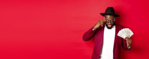 Sassy and cool Black man in hat and party outfit, showing dollars and looking from under sunglasses, standing over red background photo