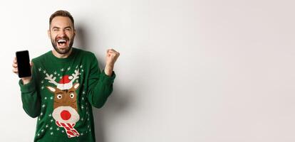 Christmas holidays and shopping concept. Excited bearded man showing mobile screen and rejoicing, winning in internet, standing over white background photo