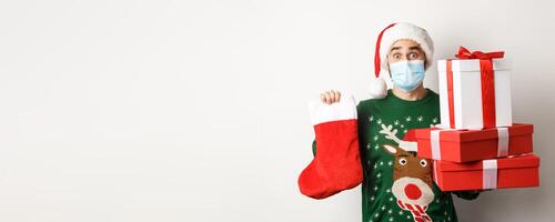Winter holidays and xmas concept. Happy man in face mask and santa hat bringing gifts, holding Christmas sock and present boxes, standing over white background photo