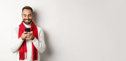 Happy man smiling while reading message on mobile phone, standing in winter sweater and red scarf against white background photo