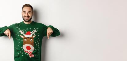 Winter holidays and christmas. Handsome smiling man with beard, pointing fingers down and showing promo offer, wearing green sweater, standing over white background photo