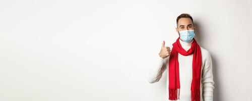 Covid-19, lockdown and quarantine concept. Satisfied smiling man in face mask showing thumb up, approve or like product, standing against white background photo