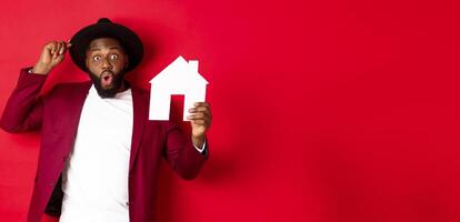Real estate. Cheerful Black man showing paper house and smiling, recommending broker, standing over red background photo