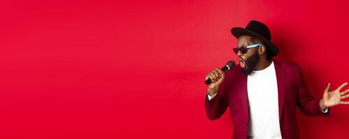 Passionate black male singer performing against red background, singing into microphone, wearing party outfit, standing over red background photo