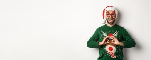 Christmas holidays, celebration concept. Happy guy in Santa hat and party glasses making fun of funny sweater, standing over white background photo