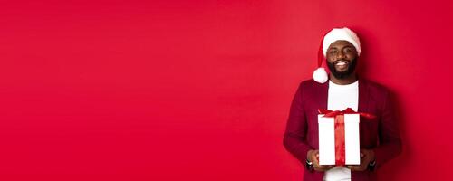 Christmas, New Year and shopping concept. Happy bearded african american man holding xmas present, smiling at camera, standing in santa hat against red background photo