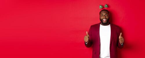 Merry Christmas. Happy african american man celebarting New Year, wearing funny party headband and showing thumb up, like and praise something, standing over red backgrond photo
