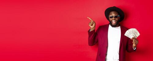 Handsome and stylish Black man pointing fingers left while showing money, holding dollars and demonstrating logo, standing over red background photo