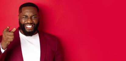 Cheerful Black man with beard wearing party clothes, pointing finger at camera and winking, congratulate with christmas and new year, standing over red background photo