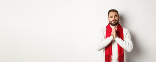 Christmas holidays and New Year concept. Desperate man begging for help, asking for favour, holding hands in pray and looking with hope at camera, standing over white background photo