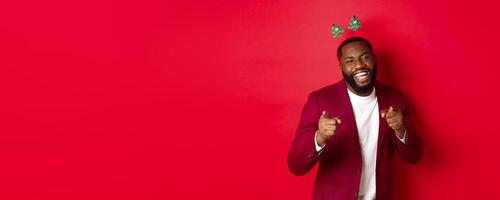 Merry Christmas. Handsome african american man in blazer and party headband, celebrating new year, pointing at camera and smiling, congratulating, red background photo