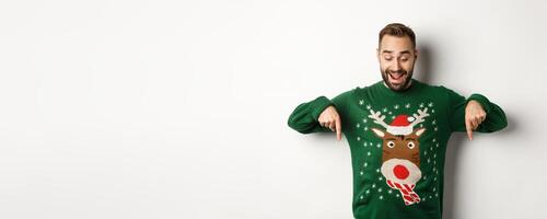 Winter holidays and christmas. Happy bearded man in green sweater, looking and pointing fingers down at advertisement, showing copy space, white background photo