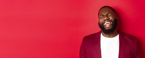 Close-up of happy and carefree Black man having fun, laughing and smiling, standing in blazer against red background photo