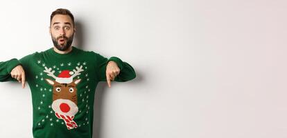 Winter holidays and christmas. Surprised bearded man in green sweater showing announcement, pointing fingers down at promo banner, white background photo