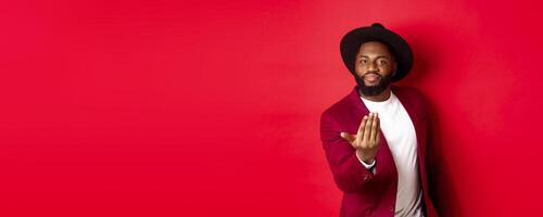 Handsome and masculine Black man asking come closer, luring to step forward, calling for you, standing over red background photo