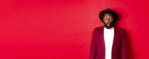 Fashion and party concept. Impressed Black man in classy outfit staring with complete disbelief at camera, gasping and looking surprised, standing over red background photo