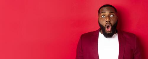 Close-up of shocked Black man gasping and dropping jaw impressed, staring at camera, standing over red background photo