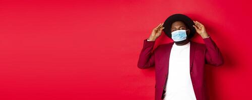 Covid-19, quarantine and holidays concept. Handsome and stylish african american man in face mask, put hat on head and looking sassy, dress-up for party, standing over red background photo